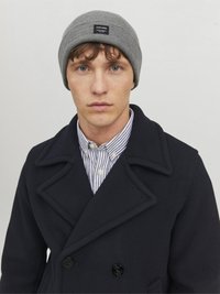 Gray ribbed beanie with a logo patch, worn with a dark navy peacoat and a striped collared shirt, featuring button details.