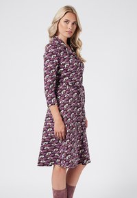 Vestido envolvente floral en púrpura con flores blancas y rosas. Presenta un lazo en la cintura, mangas 3/4 y una falda acampanada. Tejido suave y ligero.