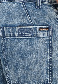 Jeans de mezclilla azul con un bolsillo trasero que lleva una etiqueta negra. La tela tiene un acabado desgastado y texturizado, con costuras dobles a lo largo de las costuras.