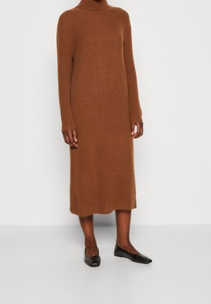 Persoon draagt een bruine, gebreide jurk met lange mouwen tot op de knieën en zwarte platte schoenen, staand tegen een effen witte achtergrond.