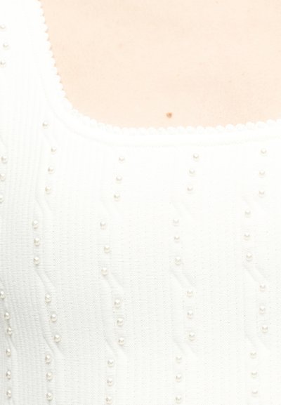 Κοντινό πλάνο λευκού πλεκτού τοπ με μικρά μαργαριταρένια χάντρες τοποθετημένες σε κάθετα σχέδια πάνω στο ύφασμα.