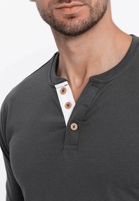 Dunkelgraues Langarmshirt mit einem weißen Einsatz und drei braunen Knöpfen. Weicher, glatter Stoff mit rundem Halsausschnitt.