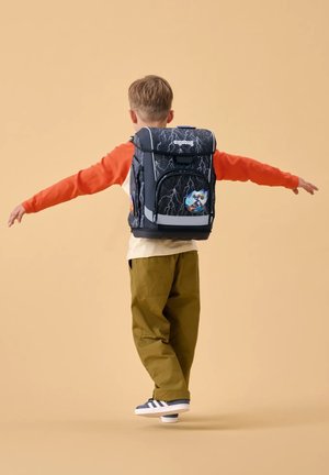 Kind balanciert auf einem Fuß mit ausgestreckten Armen, trägt orangefarbene Ärmel, olivfarbene Hose und einen schwarzen Rucksack mit Blitzdesign.