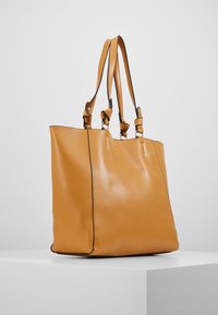 Hnědá kožená taška přes rameno s dvěma popruhy, hladkou texturou, pevný tvar a černým prošitím podél okrajů.