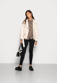Jaqueta de imitação de couro creme com fecho de zip, blusa floral, calças pretas e loafers pretos grossos. Acessórios incluem uma pequena bolsa preta.