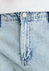 Lyseblå denimjeans med en falmet finish. Har en metallknapp med merket navn på midjebåndet og synlige sømdetaljer.