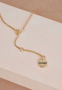 Guldkedjehalsband med cirkulär hängsmycke graverad med "Cath Kidston" och en liten infattad ädelsten, liggande på en rosa texturerad yta.