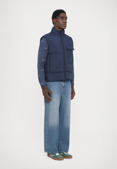 Navy blue puffer vest with high collar, front pocket, and zipper; worn over a patterned long-sleeve shirt and wide-leg denim trousers.