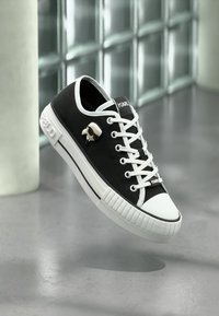 Schwarze Leinensneaker mit weißer Gummisohle, weißer Schnürung, rundem Zehenbereich und einem markanten Logo-Anstecker an der Seite.