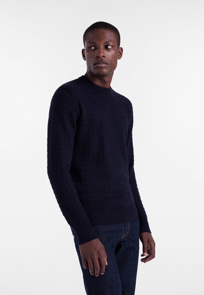 Temno modra pletena pulover s teksturiranim diamantnim vzorcem, krojaška vratna odprtina in rebrasti manšeti. Nosi se z temnimi kavbojkami.