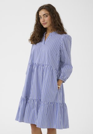 Donna con capelli ricci che indossa un vestito a righe verticali bianche e blu a maniche lunghe con orlo arricciato, in piedi con una mano in tasca.