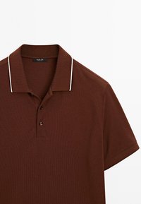Polo marron à manches courtes avec bordure blanche sur le col et patte à deux boutons, posé à plat sur un fond blanc.