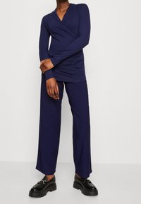 Mörkblå långärmad topp med omslagdesign och rynkade sidor, tillsammans med vida byxor. Svarta plattformsskor med dekorativa metallaccenter.