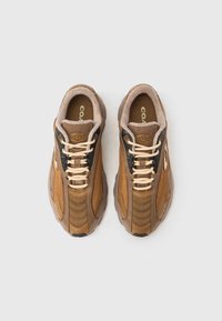 Καφέ αθλητικά παπούτσια από δίχτυ και δέρμα με υφή στην επιφάνεια, εκρού κορδόνια και επενδυμένους γιακάδες, με μαύρες λεπτομέρειες στα πλευρά και λογότυπο.