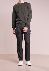 Sudadera verde oliva con cuello redondo y puños de canalé, combinada con pantalones grises oscuros con plisados y zapatillas blancas, de pie sobre un suelo claro.