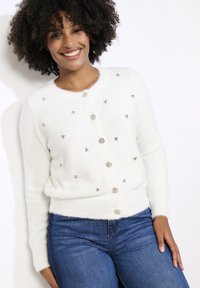 Bílý, načechraný kardigan s kulatým výstřihem, zdobený lesklými stříbrnými knoflíky a dekorativními akcenty. Spárován s modrými džíny.