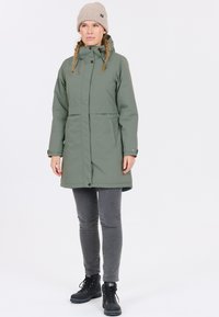 Grön vattentät jacka med huva, fickor framtill och tryckknappar, kombinerad med grå jeans och svarta snörade stövlar.