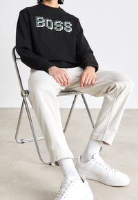 Sudadera negra con logo pixelado de "BOSS", pantalones de pana color crema y zapatillas blancas, sentado en una silla plegable metálica.