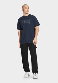 Granatowa koszulka z nadrukiem "PROSTO" w ciemnoszarym kolorze, w połączeniu z czarnymi spodniami i białymi trampkami z niebieskimi akcentami.