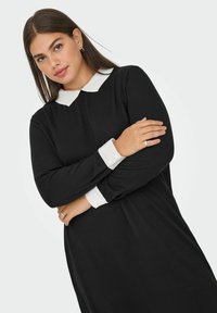 Crna haljina s dugim rukavima i bijelim ovratnikom te manšetama s gumbima. Glatki materijal s jednostavnim, opuštenim krojem.