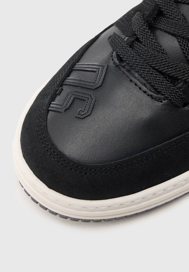 Black sneaker with a mixed material upper featuring leather and suede. Textured laces, embossed logo, and white rubber sole with subtle tread.