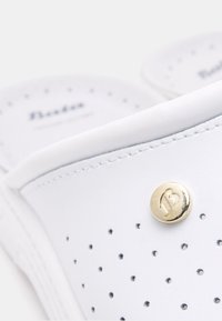 Sandales blanches à enfiler avec un design perforé, en matériau doux, et un accent de logo circulaire doré. Semelle texture pour le confort.