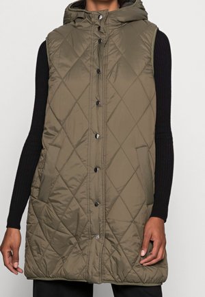Veste matelassée sans manches vert olive avec capuche et boutons-pression portée sur un haut noir à manches longues côtelé, poches visibles sur les côtés.