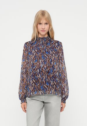 Vrouw die staat in een langhemd met abstracte patronen in blauw, bruin en beige en lichtgrijze broek tegen een witte achtergrond.