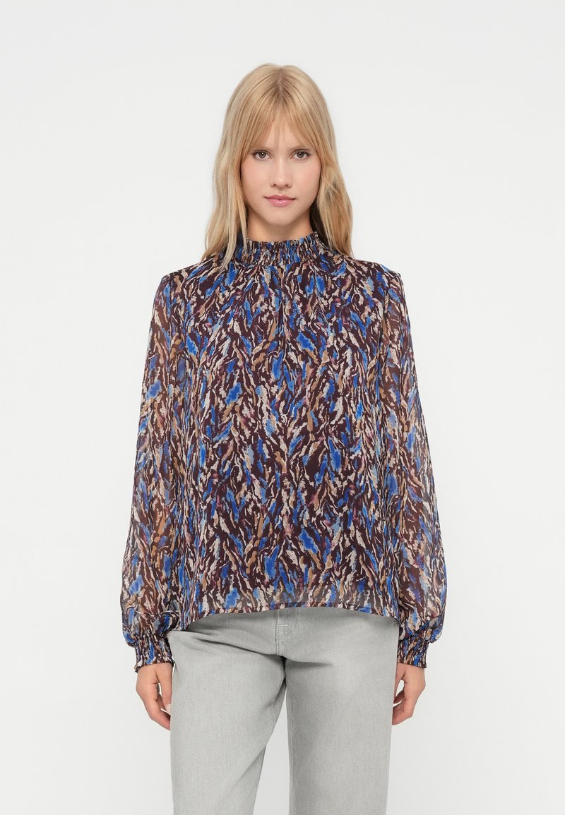 Mujer de pie con una blusa de manga larga con patrones abstractos en azul, marrón y beige y pantalones gris claro contra un fondo blanco.