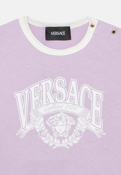 Fialové tričko s bielym logom „VERSACE“ a grafickým detailom s motívom hlavy. Okrúhly výstrih s kontrastným bielym lemom a dekoratívnymi cvokmi.