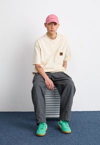 T-shirt ample beige clair avec une poche poitrine, pantalon cargo gris et baskets vertes. Le mannequin porte une casquette rose et est assis sur un siège rayé.