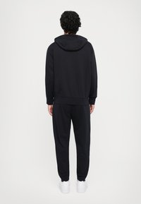 Person standing with back facing camera, wearing black hoodie and matching black sweatpants with white sneakers against a plain white background.