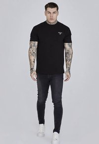 Musta lyhythihainen t-paita pyöreällä pääntiellä ja pienellä logolla rinnassa, yhdistetty kapeisiin tummiin farkkuihin ja valkoisiin lenkkareihin.