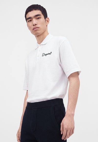 Jeune homme portant un polo blanc Dsquared2 rentré dans un pantalon noir, debout devant un fond clair uni.