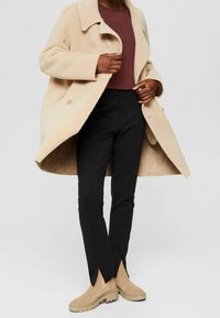 Cappotto beige e morbido con colletto a punte, due bottoni, indossato su un top bordeaux. Pantaloni neri aderenti e stivaletti beige con suola spessa.
