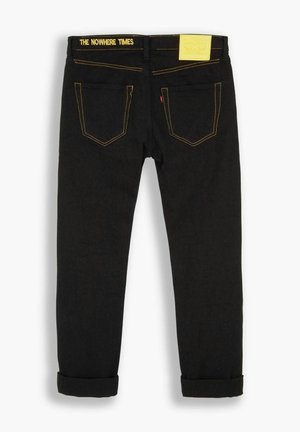 Jeans de mezclilla negra con costuras amarillas, que presentan un dobladillo enrollado, bolsillos traseros y una etiqueta de la marca en la cintura con el texto "THE NOWHERE TIMES."