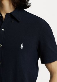 Marinblå pikétröja i texturerad tyg, med klassisk krage och vit broderad logotyp på vänster bröst, samt silverknappar.