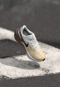 Nike trkaća cipela s bijelim mrežastim gornjim dijelom, žutim akcentima, crnim swoosh logotipom i smešim petnim dijelom, postavljena na tamnoj pozadini.