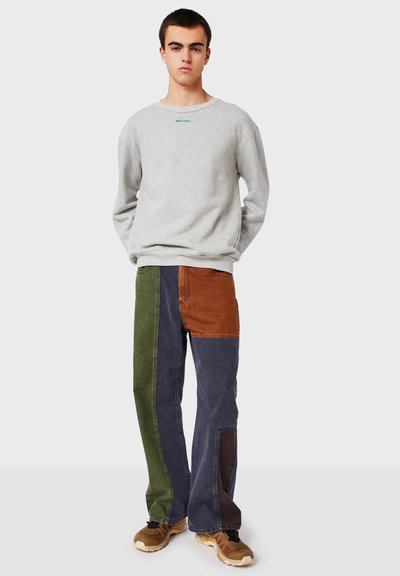 Ung man iförd en ljusgrå sweatshirt och byxor i lapptäcke med gröna, bruna och blå paneler, stående mot en enhetlig bakgrund.