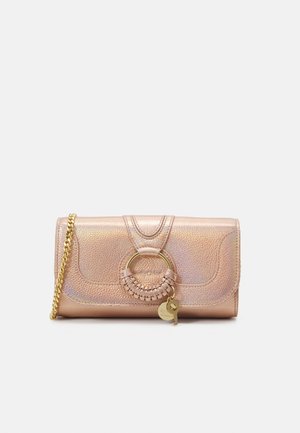 Petit sac à main en cuir métallisé rose doré avec bandoulière chaîne dorée, détail anneau tressé circulaire et deux petits pendentifs charme dorés.