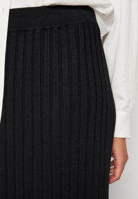 Černá, řasená plisovaná sukně s elastickým pasem. Tkanina vypadá texturovaně a zobrazuje vertikální pruhy po délce.
