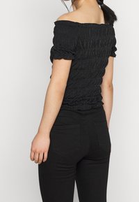 Svart off-shoulder topp med smockad textur, korta puffärmar och volangkant. Kombinerad med svarta tajta jeans.