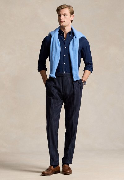 Mężczyzna stoi z rękami w kieszeniach, ubrany w granatową koszulę, jasnoniebieski sweter zarzucony na ramiona, spodnie w prążki i brązowe buty.