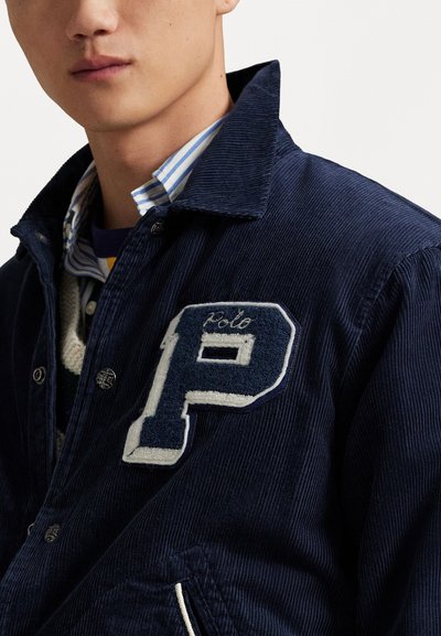 Navy corduroy jacket featuring a textured 'P' patch, snap buttons, and side pockets, layered over a striped shirt.