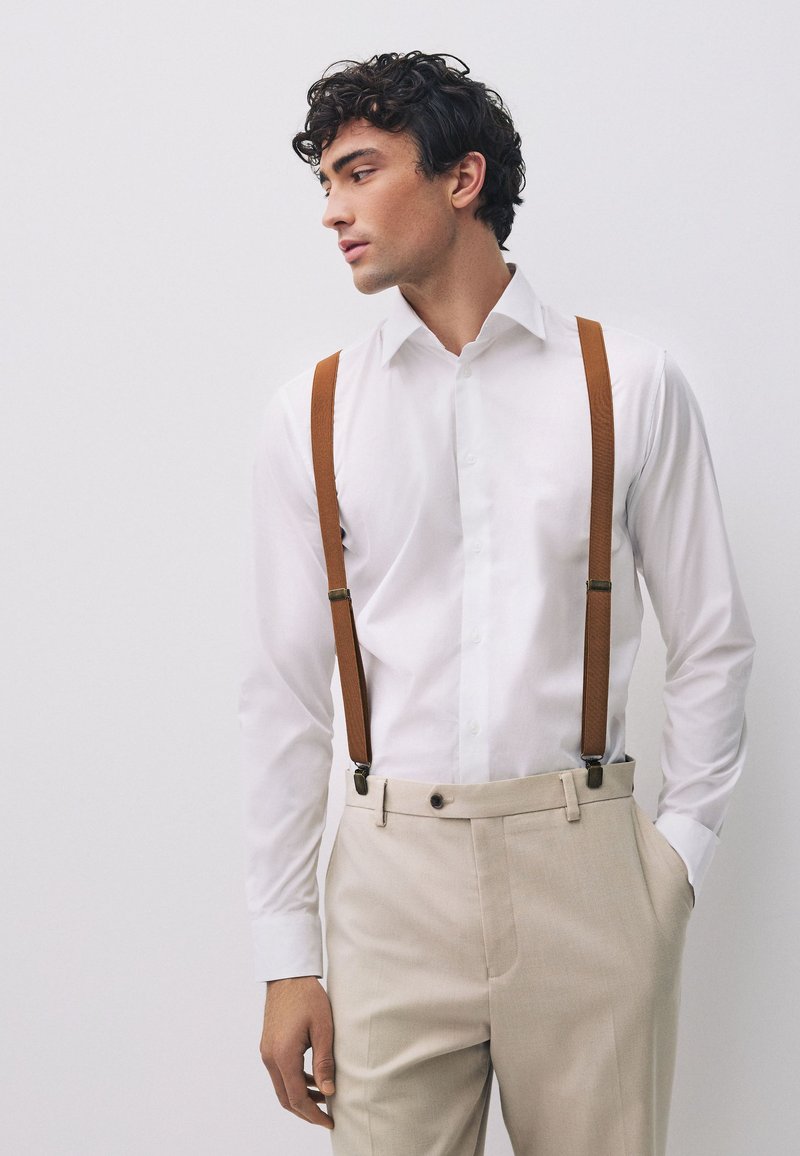 Giovane uomo con camicia bianca, pantaloni beige e bretelle marroni, guarda a sinistra con una mano in tasca davanti a uno sfondo semplice.