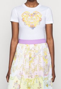Biała bawełniana koszulka z ozdobnym żółto-niebieskim motywem serca, w połączeniu z plisowaną spódnicą w pastelowych kolorach i kwiatowymi wzorami.