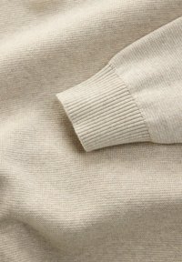 Beige gebreide trui mouw met geribde manchet op een bijpassende stof achtergrond, die textuur en fijne brei-details toont.