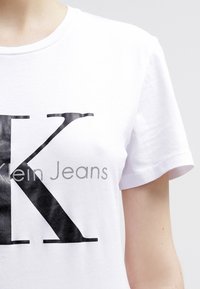 Vit t-shirt i bomull med en stor svart "K"-logotyp och grå text "lein Jeans" tryckt nedanför, med rund halsringning och korta ärmar.