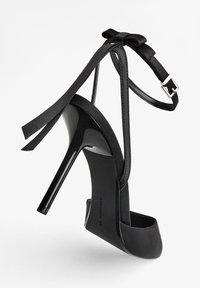 Sandal con tacco alto nera caratterizzata da una piattaforma in pelle verniciata elegante, sottili fasce alla caviglia e un accento in satin a forma di fiocco sul retro.