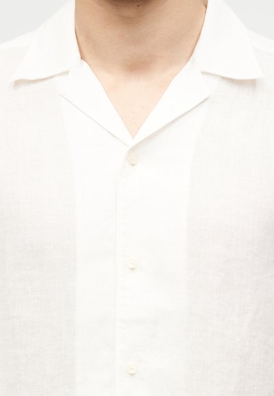 Άνδρας που φοράει ένα λευκό πουκάμισο με κουμπιά, ελαφρώς ανοιχτό, με χαλαρό γιακά, που αποκαλύπτει μέρος του λαιμού και του άνω μέρους του στήθους.